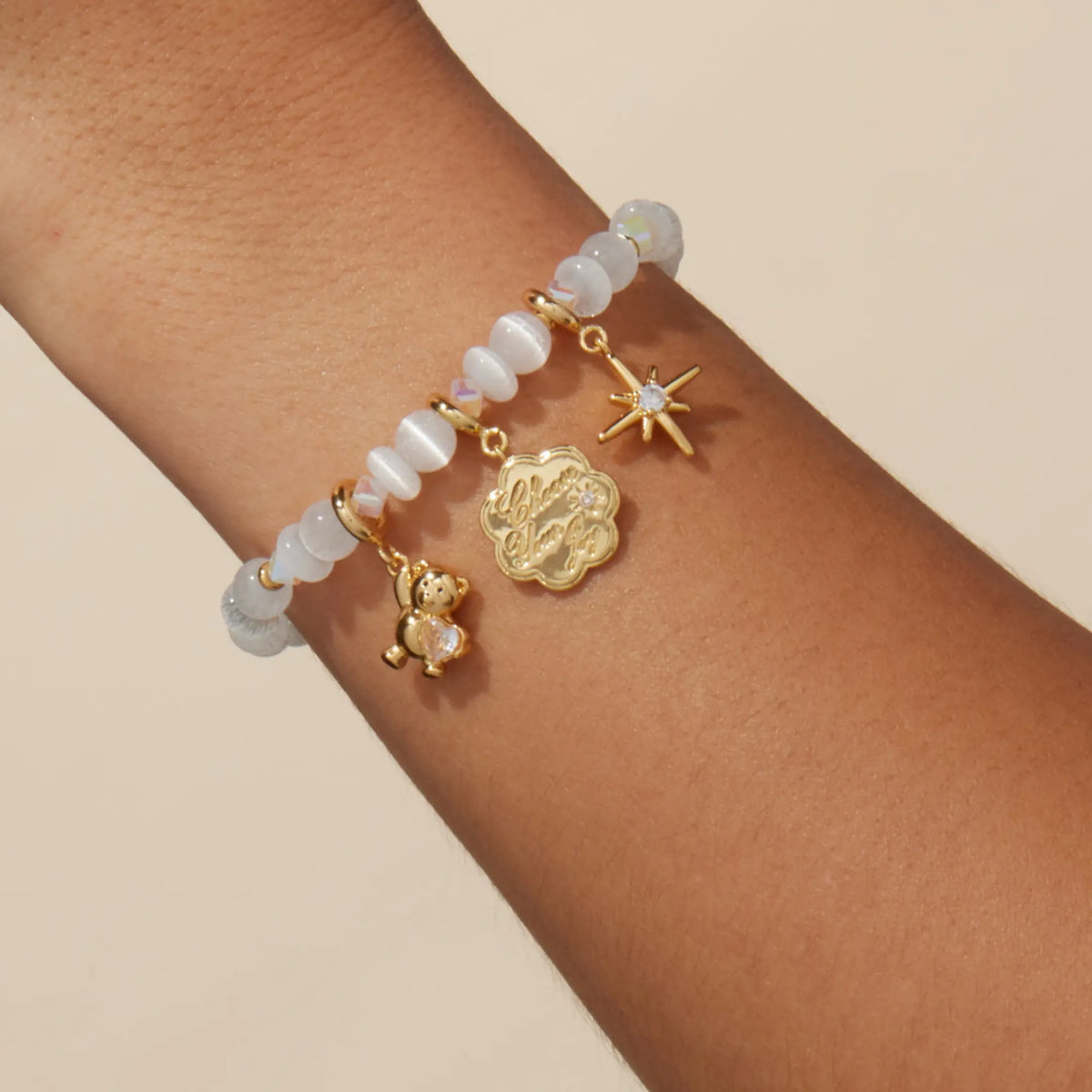 Arctic Honey Bracelet Base worn on a wrist styled with gold teddy bear gem, Choose Your Joy coin, and starburst charms, on-model on a tan background