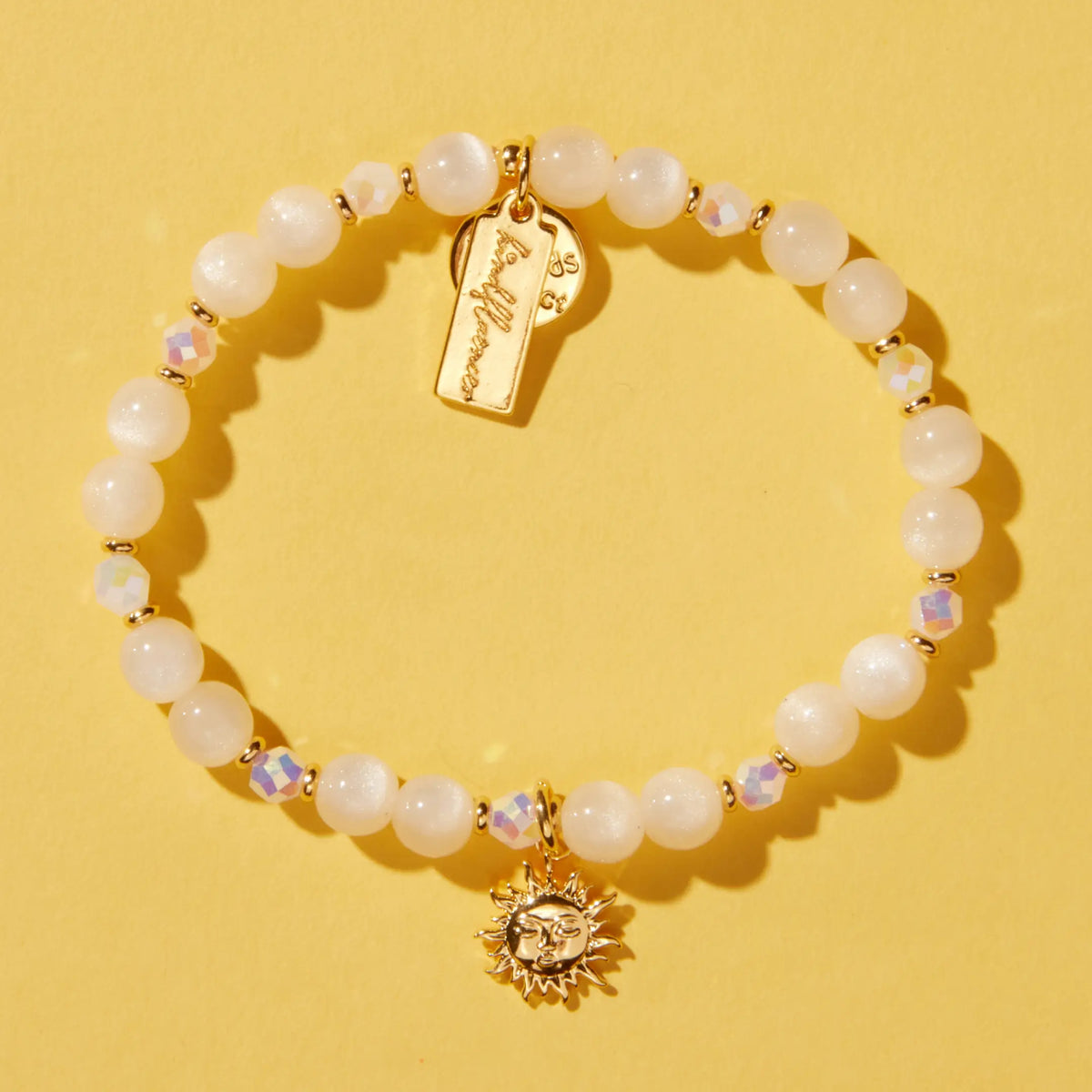 Sunshine Bracelet Base by Anna Sitar, white resin cat eye and iridescent crystal beads with a gold sun charm and gold Kindfluencer tag, flat-lay on yellow background