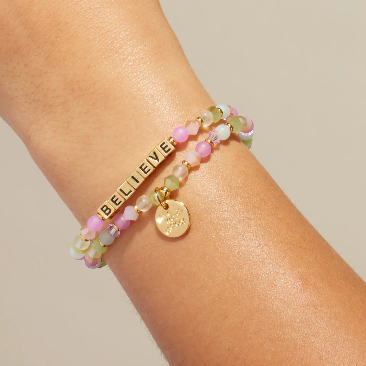 Gold Believe bracelet in the Butterfly Effect bead pattern with gold letter beads, stacked with a coordinating beaded bracelet, on-model on wrist against cream background