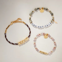 Lucky Charm Set flat-lay on cream background showing three stacked bracelets: BOLDER Lucky paracord bracelet, Trust bracelet in gold letter beads, and You Got This dalmatian jasper bracelet