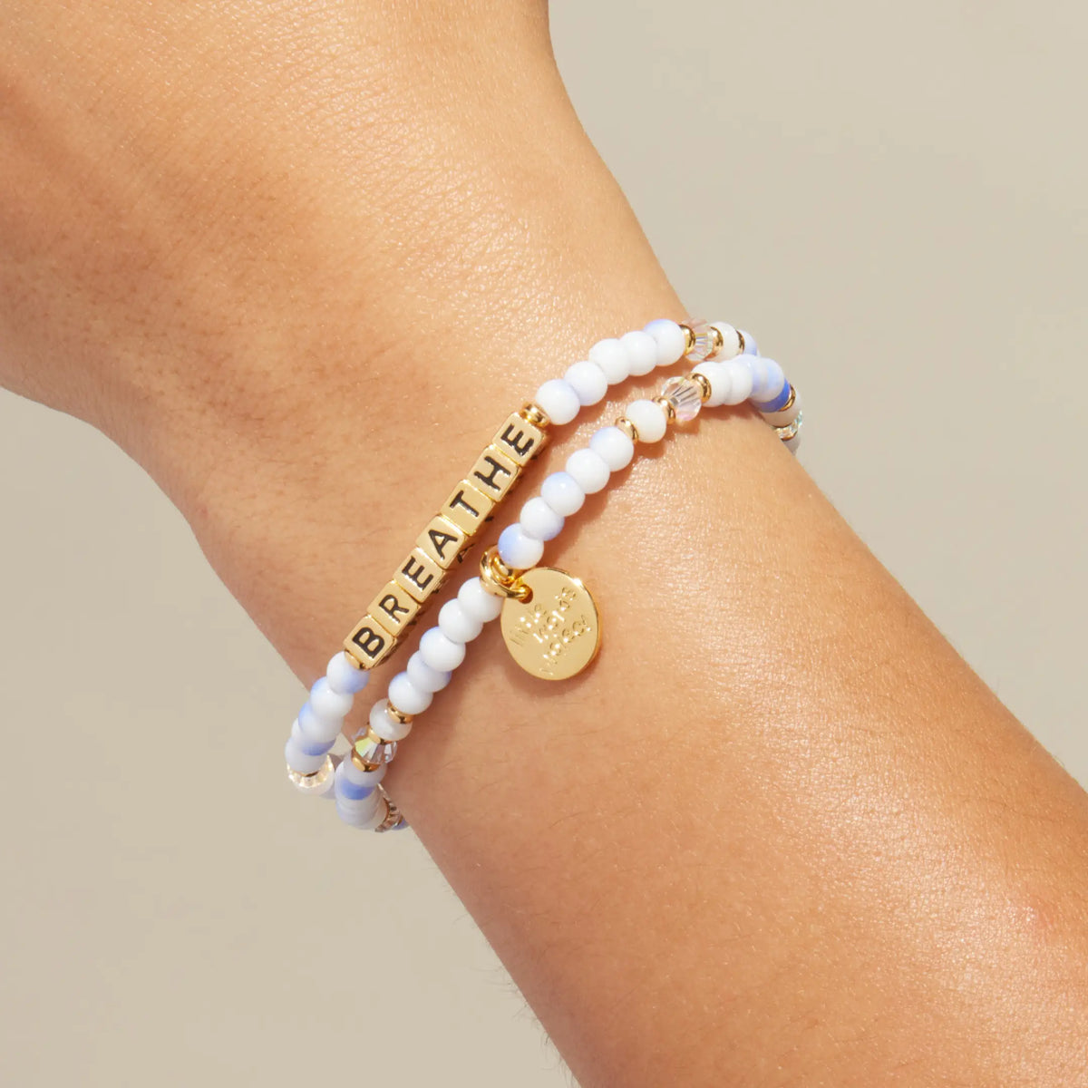 Gold Breathe bracelet in the Cotton Clouds bead pattern with gold letter beads, stacked with a coordinating beaded bracelet, on-model on wrist against cream background