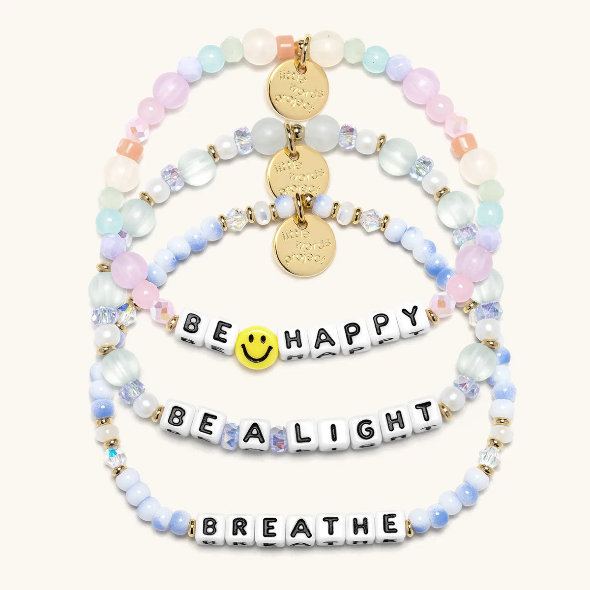 Good as Gold Bracelet Set in white letter beads, flat-lay on cream background showing three stacked bracelets: Be Happy with a yellow smiley face bead and pastel beads, Be a Light in seafoam glass and pearl beads, and Breathe in blue and white swirl beads