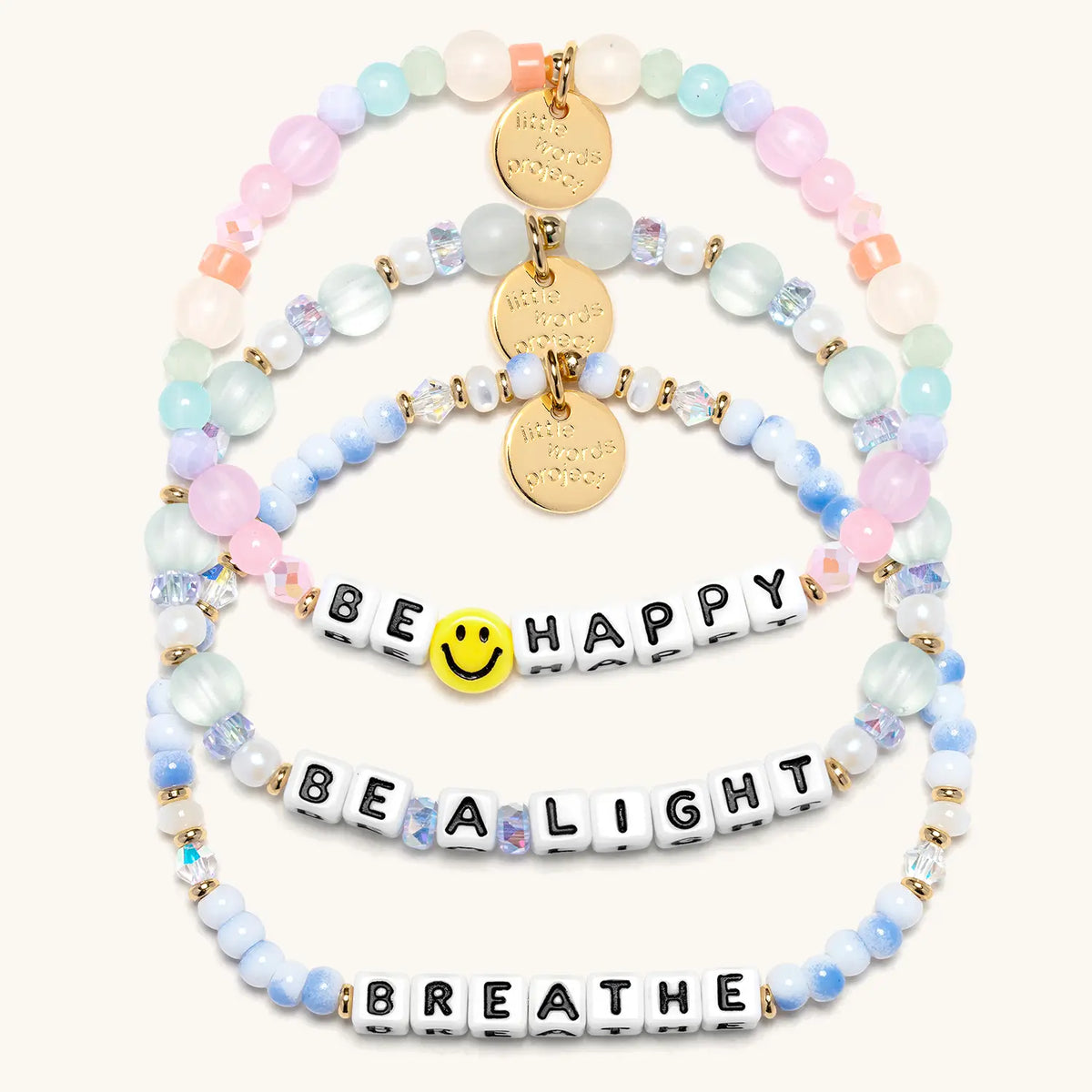 Good as Gold Bracelet Set in white letter beads, flat-lay on cream background showing three stacked bracelets: Be Happy with a yellow smiley face bead and pastel beads, Be a Light in seafoam glass and pearl beads, and Breathe in blue and white swirl beads