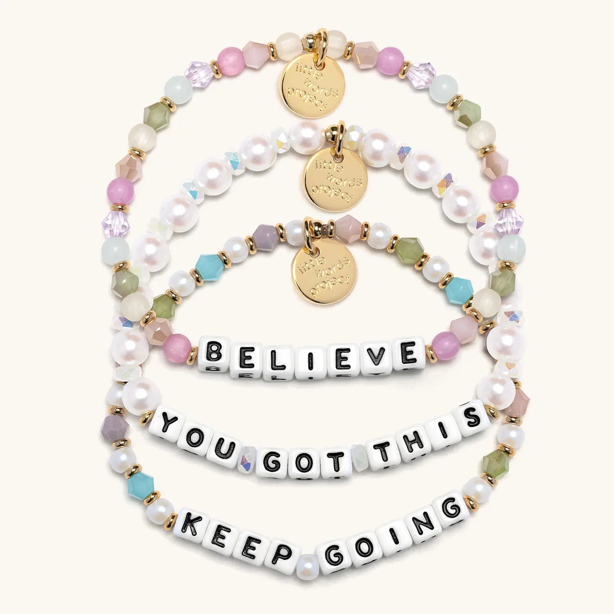 Good Things Ahead Bracelet Set in white letter beads, flat-lay on cream background showing three stacked bracelets: Believe in orchid, mint and blush beads, You Got This in white faux pearls and iridescent crystals, and Keep Going in pearls and jewel tone crystals