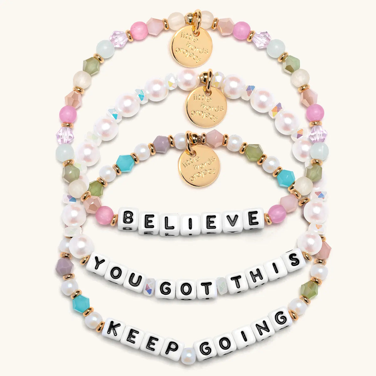 Good Things Ahead Bracelet Set in white letter beads, flat-lay on cream background showing three stacked bracelets: Believe in orchid, mint and blush beads, You Got This in white faux pearls and iridescent crystals, and Keep Going in pearls and jewel tone crystals