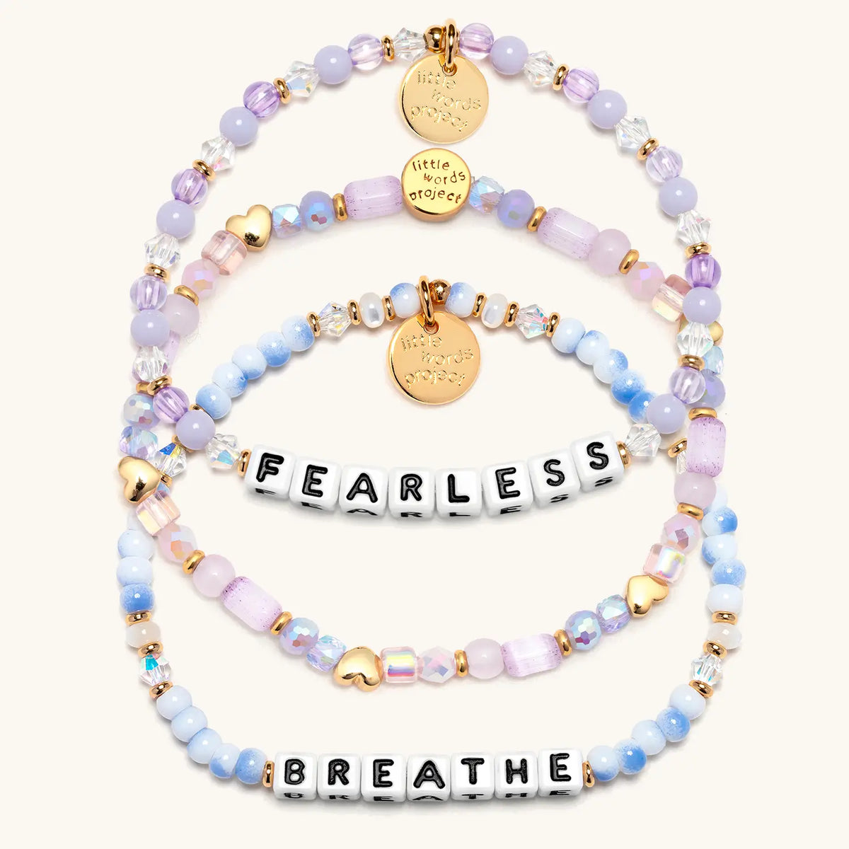 Soft & Strong Bracelet Set in white letter beads, alternate flat-lay on cream background showing the Hearts by the Yard stacker with gold heart beads, Fearless in lavender beads and crystals, and Breathe in blue and white swirl beads