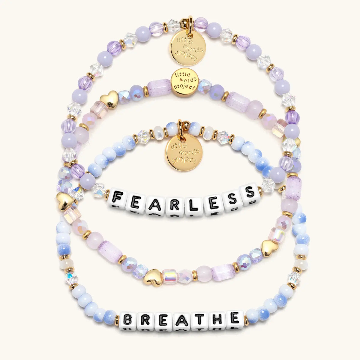 Soft & Strong Bracelet Set in white letter beads, flat-lay on cream background showing three stacked bracelets: Fearless in lavender beads and crystals, the Hearts by the Yard stacker with gold heart beads, and Breathe in blue and white swirl beads