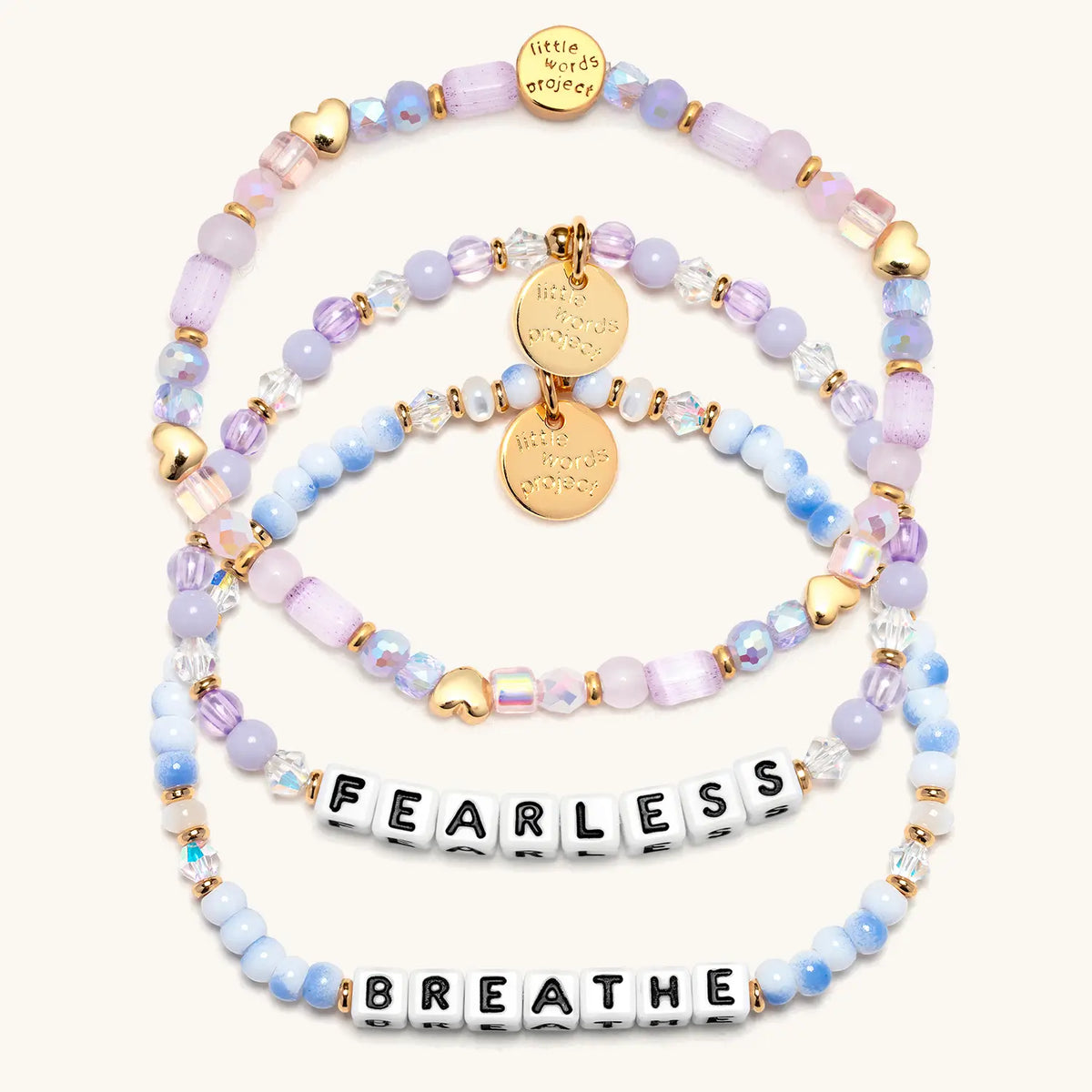 Soft & Strong Bracelet Set in white letter beads, flat-lay on cream background showing three stacked bracelets: Fearless in lavender beads and crystals, the Hearts by the Yard stacker with gold heart beads, and Breathe in blue and white swirl beads