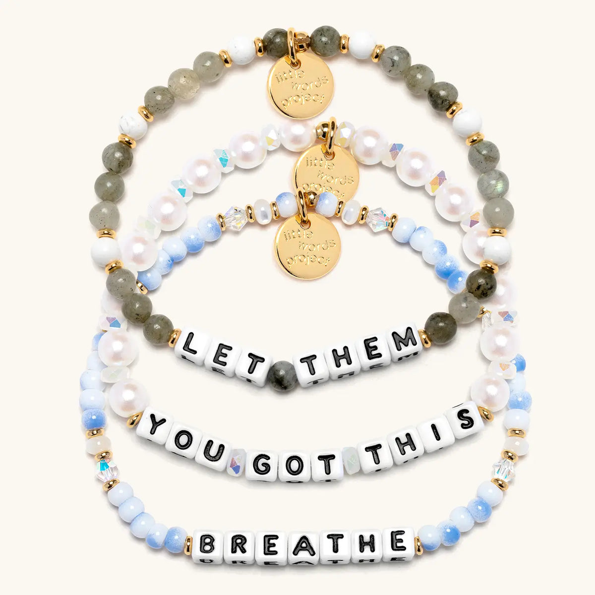 The Reset Bracelet Set in white letter beads, flat-lay on cream background showing three stacked bracelets: Let Them in gray and white stone beads, You Got This in white faux pearls and iridescent crystals, and Breathe in blue and white swirl beads