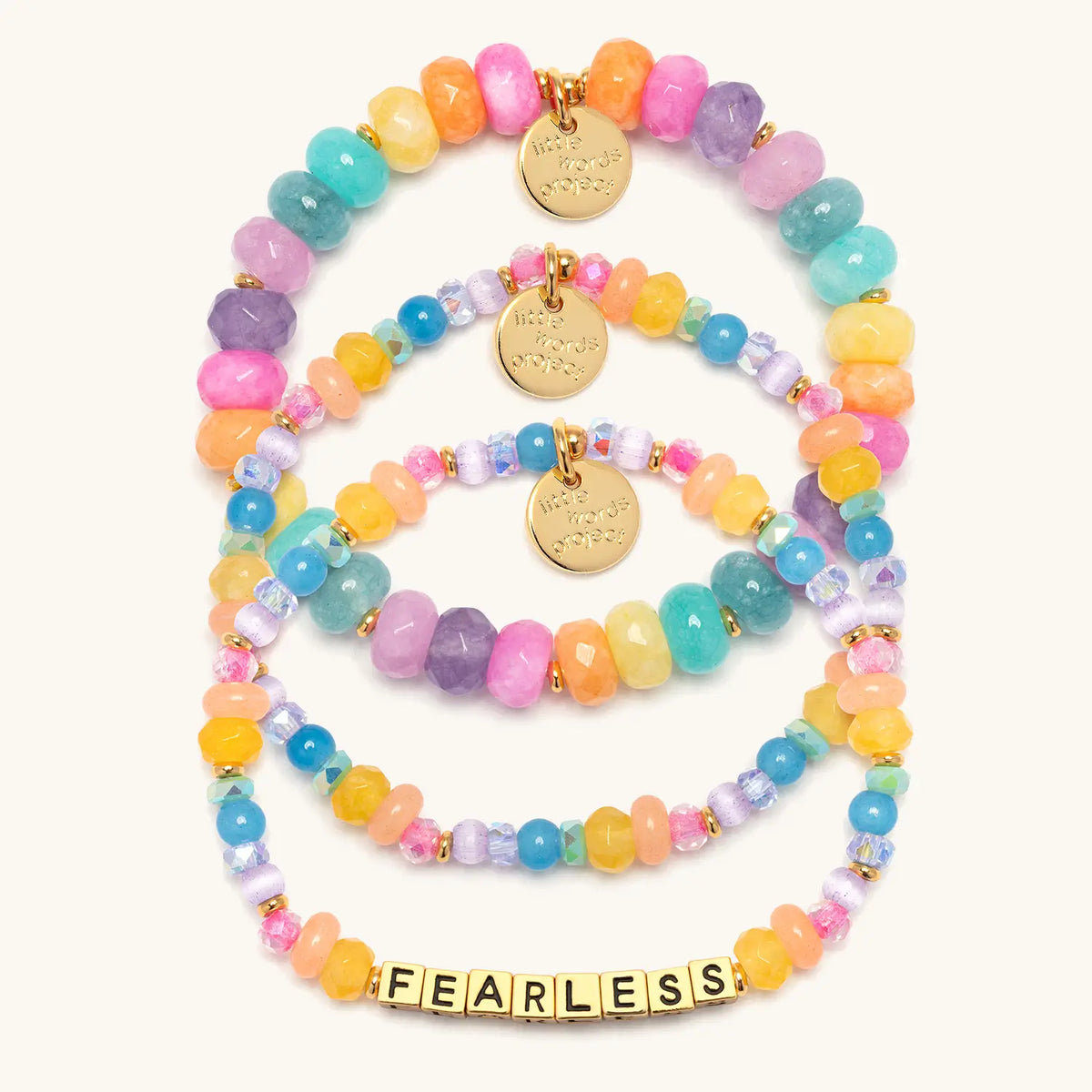 Fearless Bracelet Set- Britt Horton flat-lay on cream background showing three stacked bracelets: the Fearless bracelet in gold letter beads with Gemstone Glow beads, the Jewel Party Bracelet Base in blue, yellow, and lavender, and the Candy Confetti Bracelet Base in rainbow faceted semi precious stones