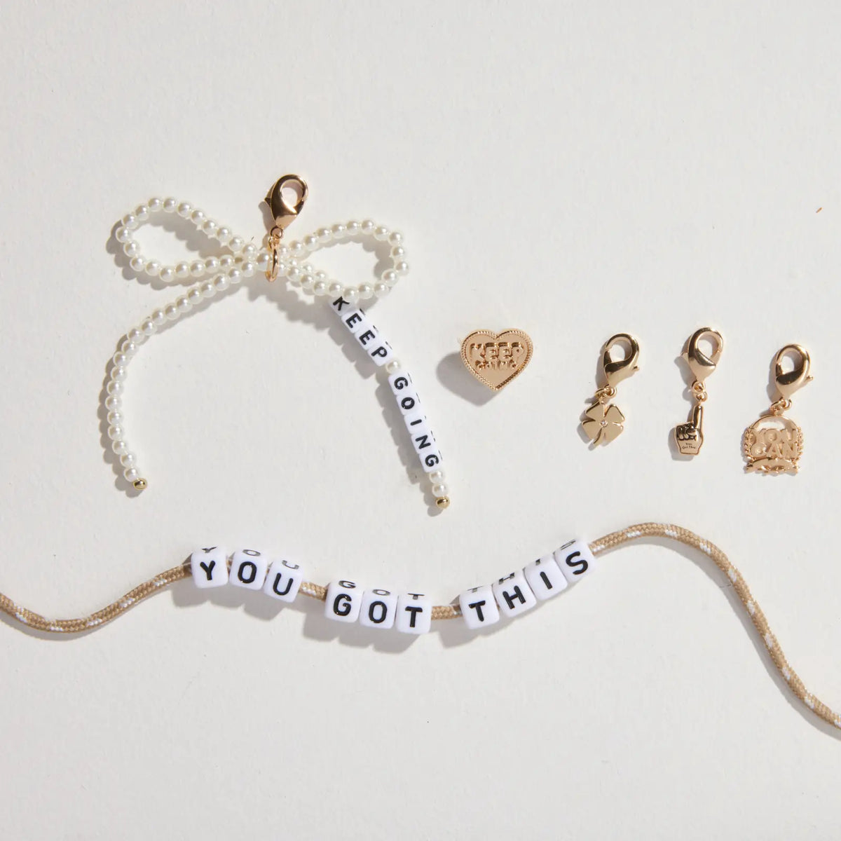 Shoe Lace Charm Kit contents laid out: white pearl beaded bow charm with Keep Going letter beads, gold Keep Going heart pin, gold four leaf clover, foam finger, and You Can charms, and tan shoelace with You Got This letter beads, flat-lay on white background