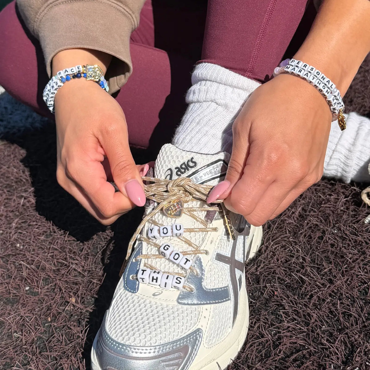 Shoe Lace Charm Kit on-model, runner lacing a cream and silver running sneaker with tan shoelaces strung with You Got This letter beads and a gold Keep Going heart pin, wearing Little Words Project bracelets on a track