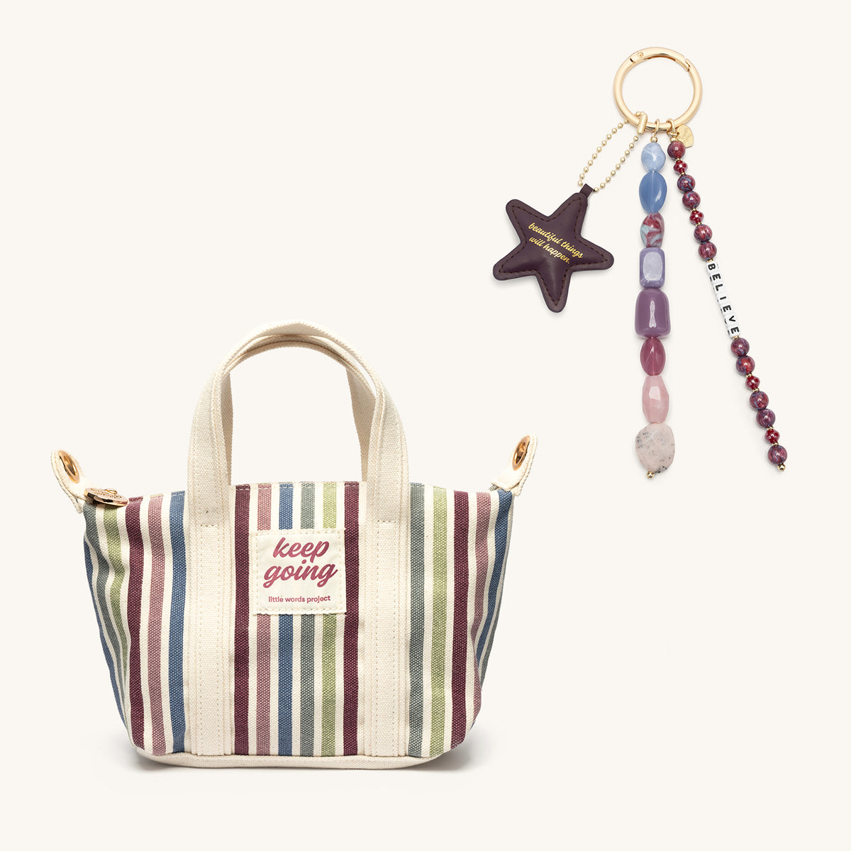Beautiful Things Set, striped Keep Going Mini Tote shown with the Beautiful Things Can Happen Bag Charm, plum faux leather star and jewel-toned beaded strands with a Believe letter strand, flat-lay on cream background