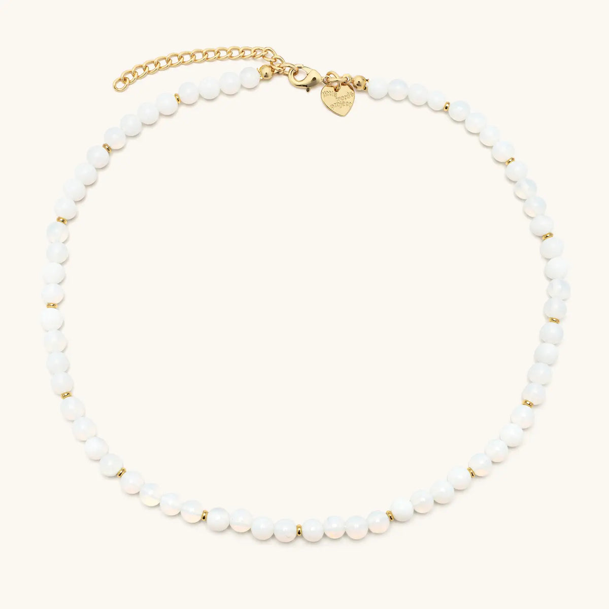 Opal Light Necklace laid flat in a circle, glowing white beads with gold spacers, gold extender chain, and gold heart logo tag, flat-lay on cream background