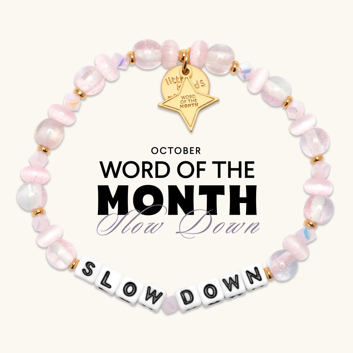 Slow Down October Word of the Month bracelet with white letter beads, pale pink glitter glass, cat eye, and iridescent crystal beads, gold spacers, and a gold star Word of the Month charm, flat-lay graphic on cream background