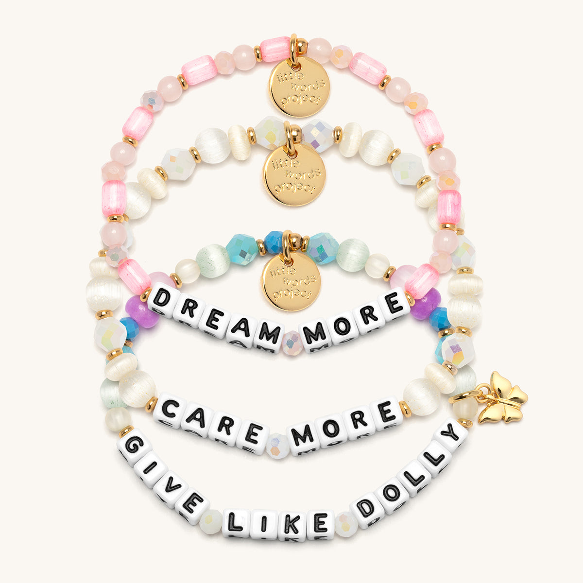 Give Like Dolly Bracelet Set flat-lay on cream background showing three stacked bracelets: the Dream More bracelet in pink tube beads, rose quartz, and blush crystals, the Care More bracelet in ivory cat's eye beads and white AB crystals, and the Give Like Dolly bracelet in aqua, pink, and purple beads with a gold butterfly charm