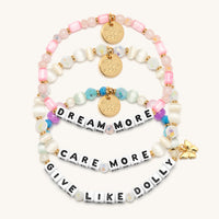 Give Like Dolly Bracelet Set flat-lay on cream background showing three stacked bracelets: the Dream More bracelet in pink tube beads, rose quartz, and blush crystals, the Care More bracelet in ivory cat's eye beads and white AB crystals, and the Give Like Dolly bracelet in aqua, pink, and purple beads with a gold butterfly charm