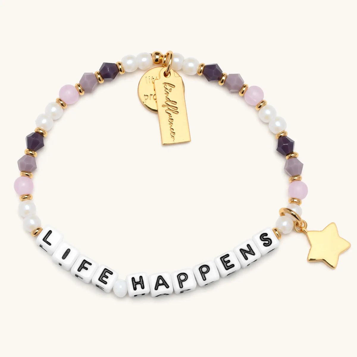 A single beaded bracelet spelling "LIFE HAPPENS" in white square letter beads with black text. The bracelet features a mix of white pearl beads, soft pink rose quartz, and deep purple amethyst-style faceted beads, with small gold spacers throughout. Two charms hang from the bracelet — a gold rectangular tag with cursive branding and a small gold star charm — displayed on a white background.