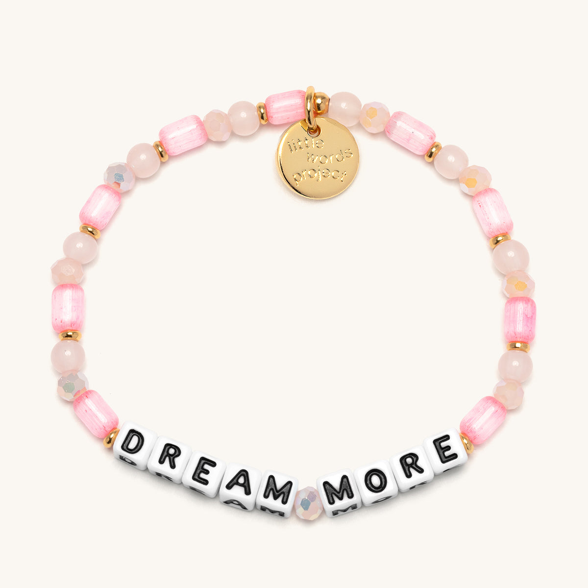Dream More bracelet with white letter beads, pink glass tube beads, rose quartz rounds, and blush AB faceted crystals with gold accents, flat-lay on cream background