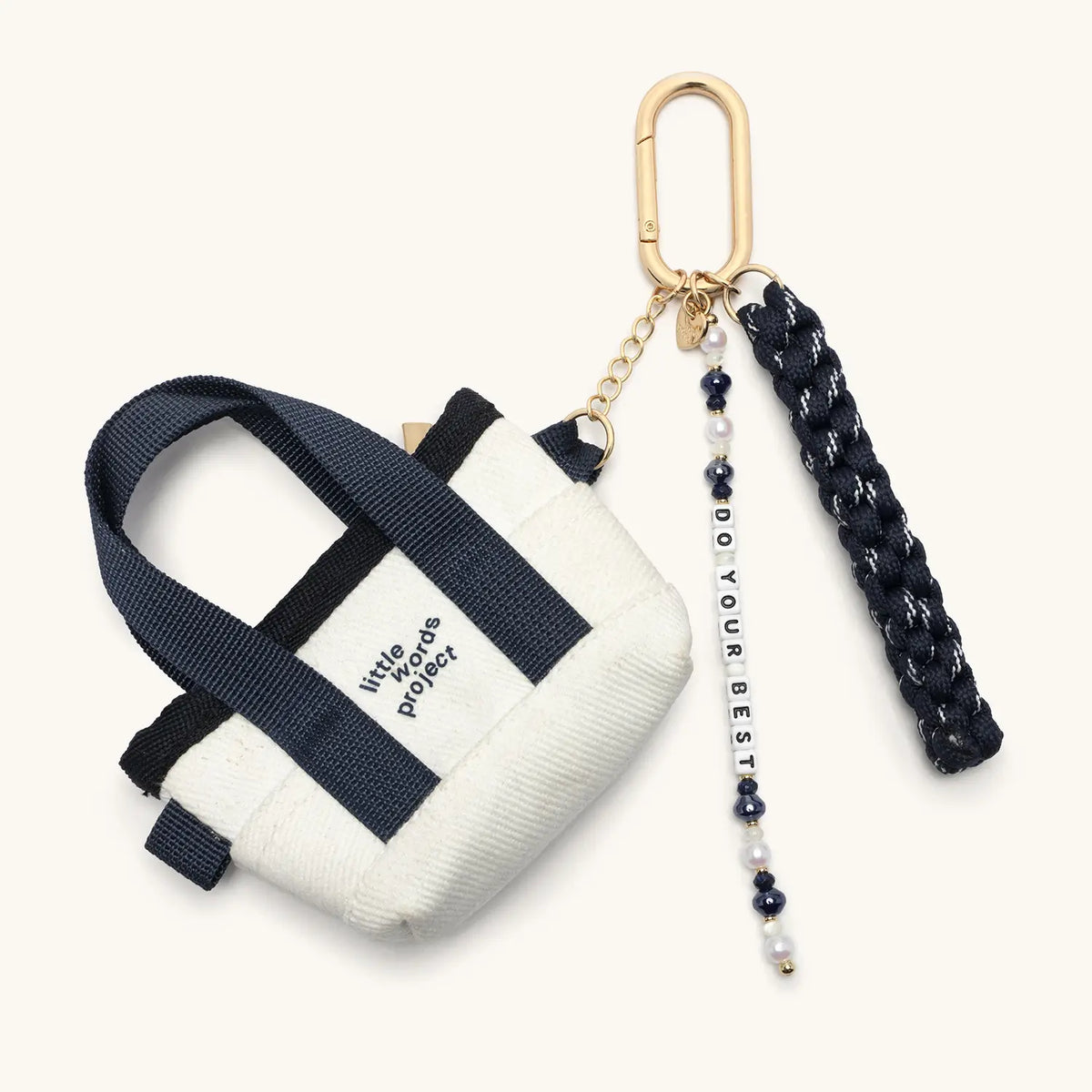 Do Your Best Bag Charm, mini cream canvas tote with navy straps and little words project logo, navy paracord tassel, and pearl and navy beaded Do Your Best word strand on a gold clip, flat-lay on cream background