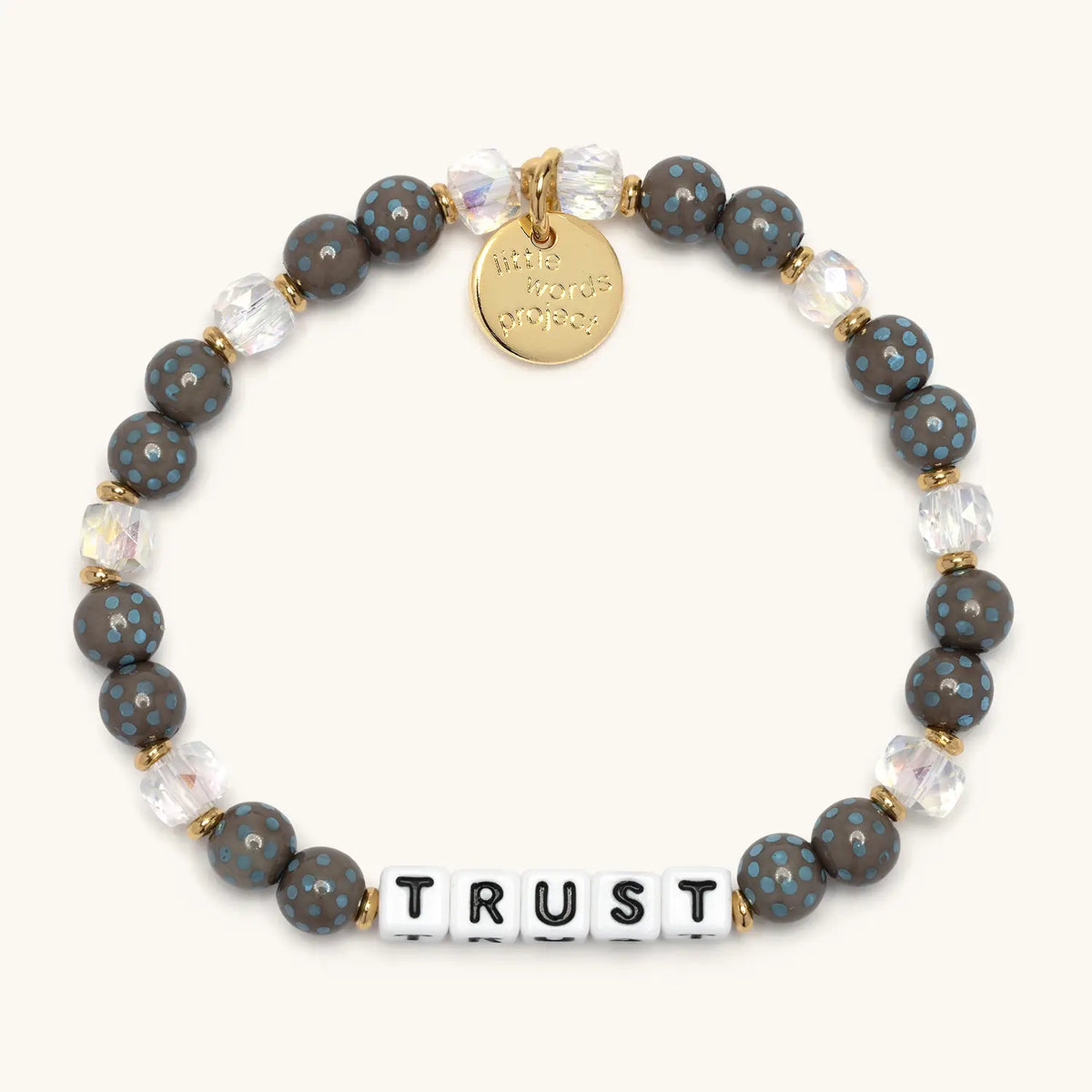 Trust bracelet with white letter beads in the Polky Babe bead pattern, dark gray beads with blue polka dots and clear iridescent crystal beads with gold accents, flat-lay on cream background