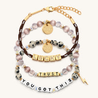 Lucky Charm Set flat-lay on cream background showing three stacked bracelets: the BOLDER Lucky paracord bracelet in brown with gold CZ letter blocks, the Trust bracelet in gold letter beads with mauve satin and clear crystal beads, and the You Got This bracelet in white letter beads with dalmatian jasper beads and a gold lightning bolt charm