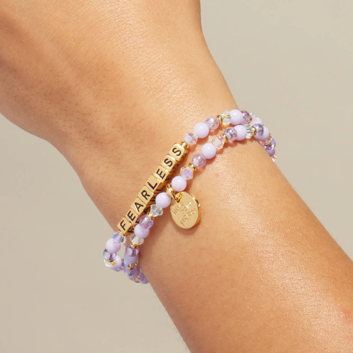 Gold Fearless bracelet in the Purple Haze bead pattern with gold letter beads, stacked with a coordinating beaded bracelet, on-model on wrist against cream background