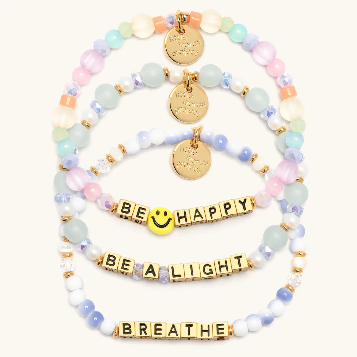 Good as Gold Bracelet Set in gold letter beads, flat-lay on cream background showing three stacked bracelets: Be Happy with a yellow smiley face bead and pastel beads, Be a Light in seafoam glass and pearl beads, and Breathe in blue and white swirl beads