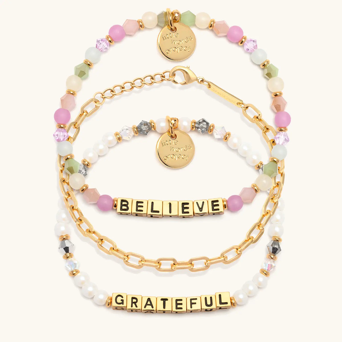 Little Reminders Bracelet Set in gold letter beads, alternate flat-lay on cream background showing Believe in orchid, mint and blush beads layered with the 14k gold-plated Paperclip Bracelet Base, and Grateful in white faux pearls and smoky crystals