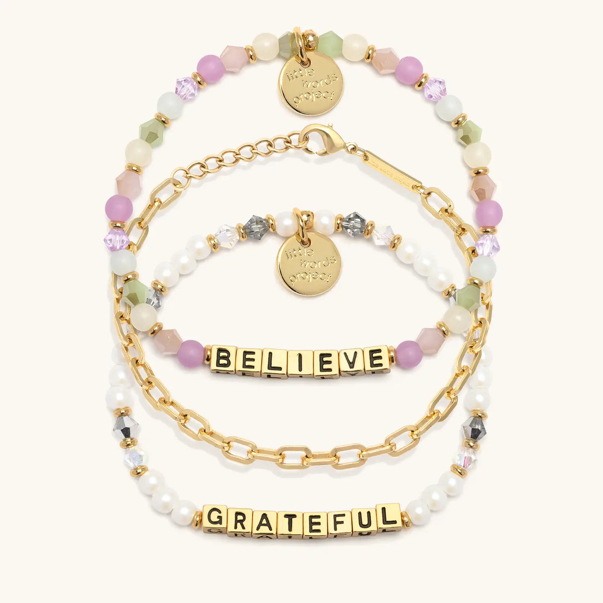 Little Reminders Bracelet Set in gold letter beads, flat-lay on cream background showing Believe in orchid, mint and blush beads, the 14k gold-plated Paperclip Bracelet Base, and Grateful in white faux pearls and smoky crystals