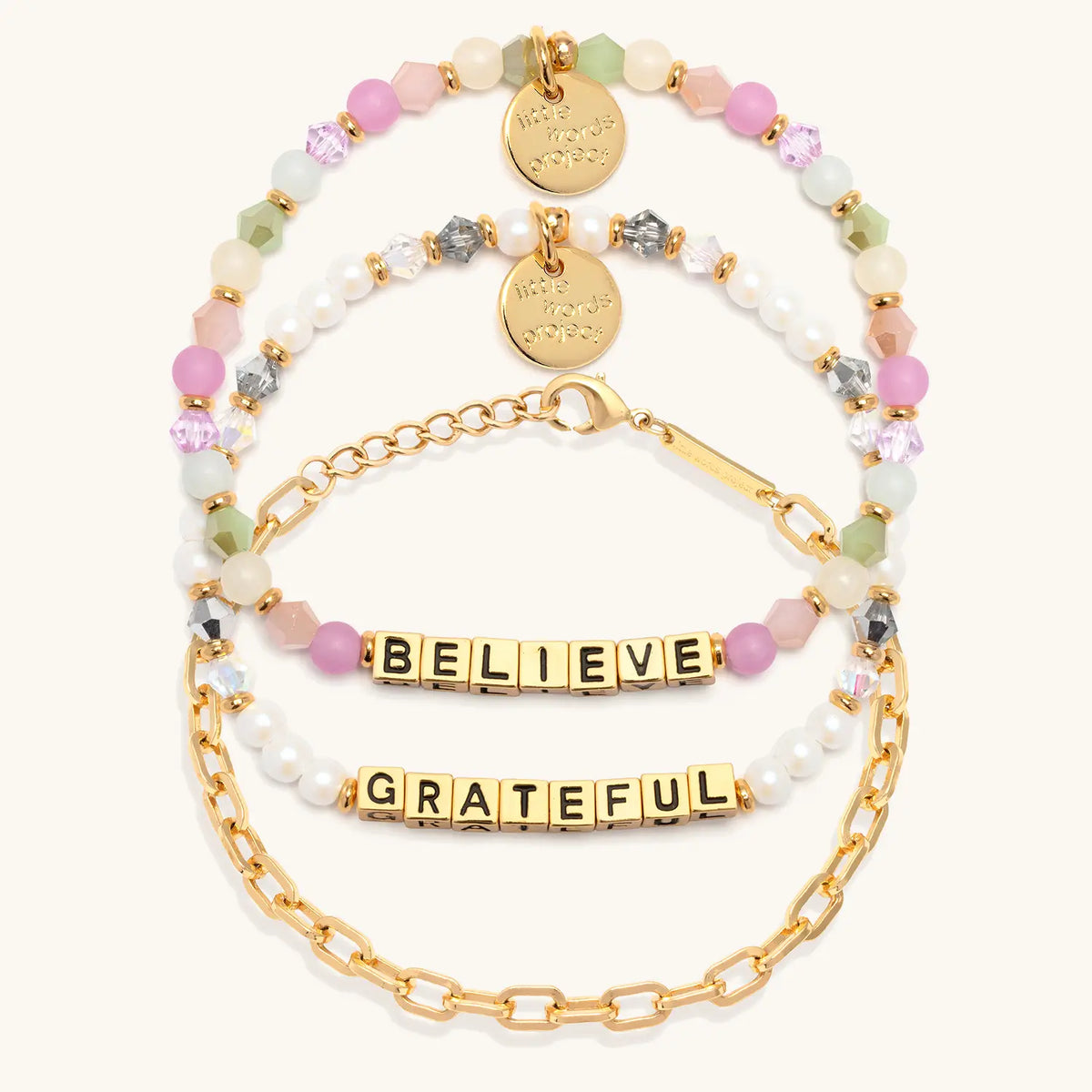 Little Reminders Bracelet Set in gold letter beads, flat-lay on cream background showing Believe in orchid, mint and blush beads, the 14k gold-plated Paperclip Bracelet Base, and Grateful in white faux pearls and smoky crystals