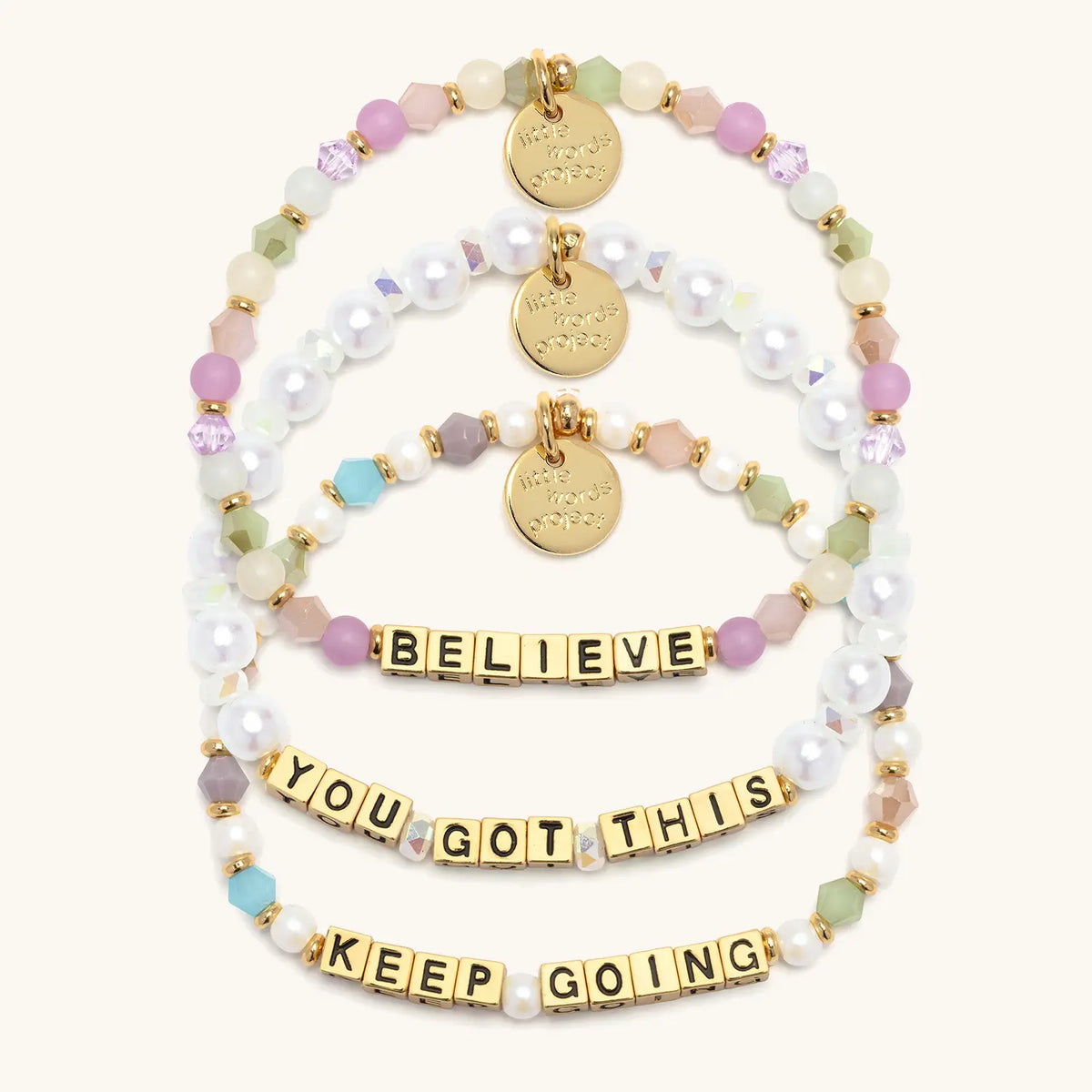 Good Things Ahead Bracelet Set in gold letter beads, flat-lay on cream background showing three stacked bracelets: Believe in orchid, mint and blush beads, You Got This in white faux pearls and iridescent crystals, and Keep Going in pearls and jewel tone crystals