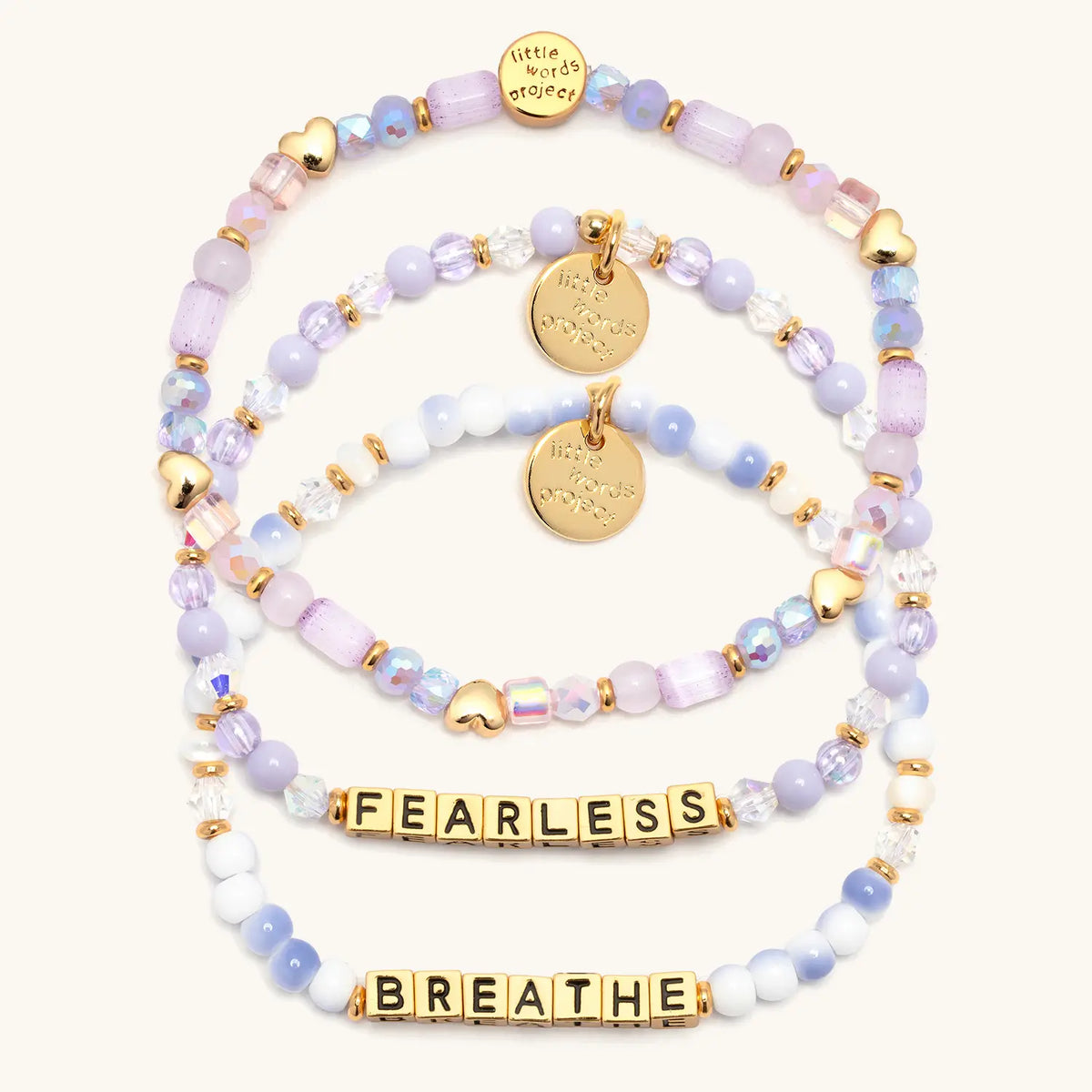 Soft & Strong Bracelet Set in gold letter beads, flat-lay on cream background showing three stacked bracelets: Fearless in lavender beads and crystals, the Hearts by the Yard stacker with gold heart beads, and Breathe in blue and white swirl beads