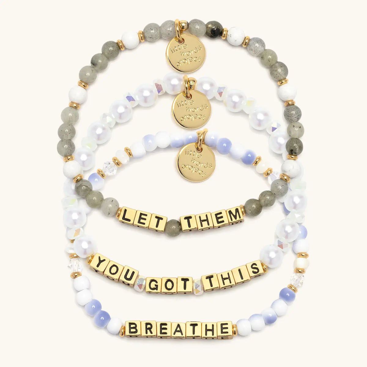 The Reset Bracelet Set in gold letter beads, flat-lay on cream background showing three stacked bracelets: Let Them in gray and white stone beads, You Got This in white faux pearls and iridescent crystals, and Breathe in blue and white swirl beads