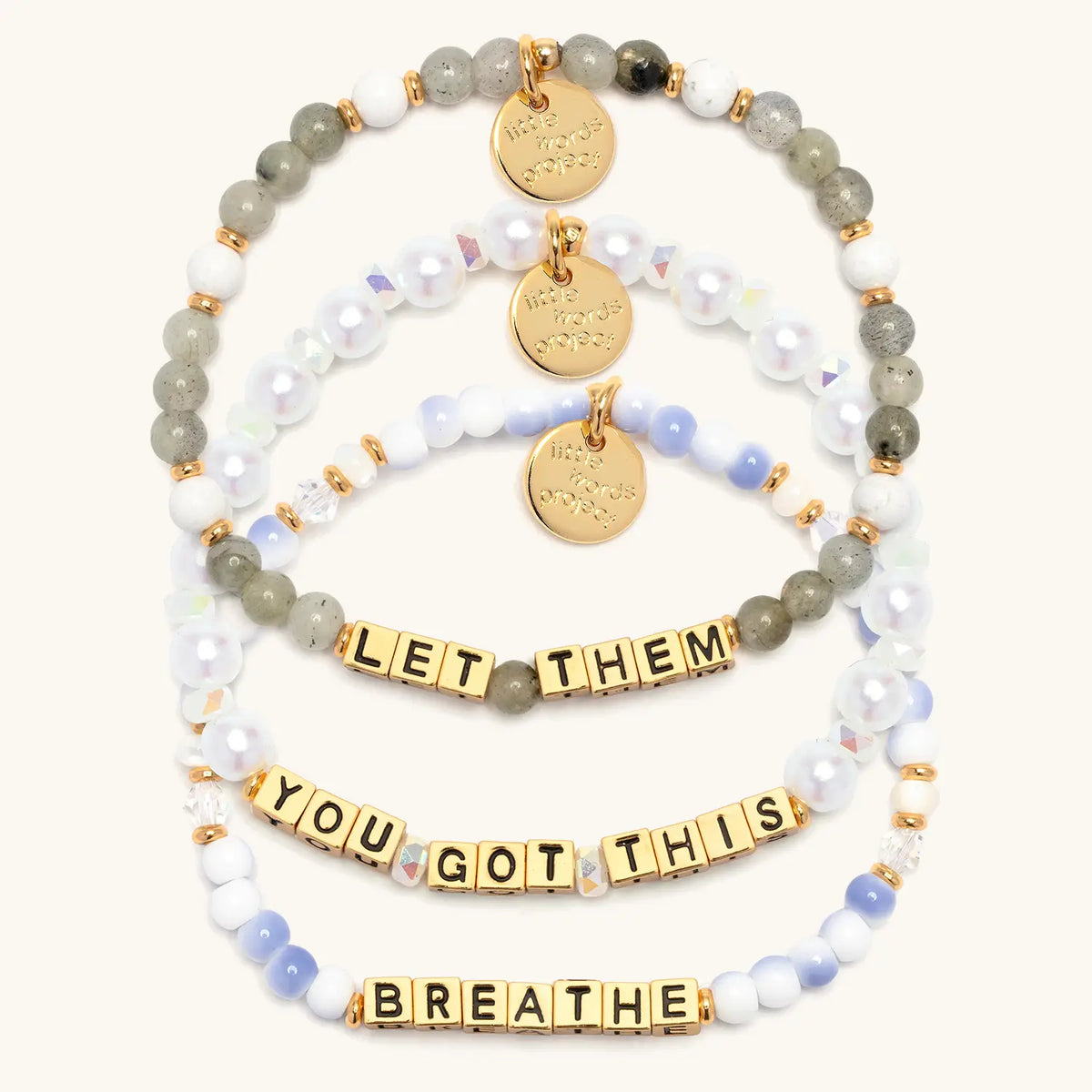 The Reset Bracelet Set in gold letter beads, flat-lay on cream background showing three stacked bracelets: Let Them in gray and white stone beads, You Got This in white faux pearls and iridescent crystals, and Breathe in blue and white swirl beads