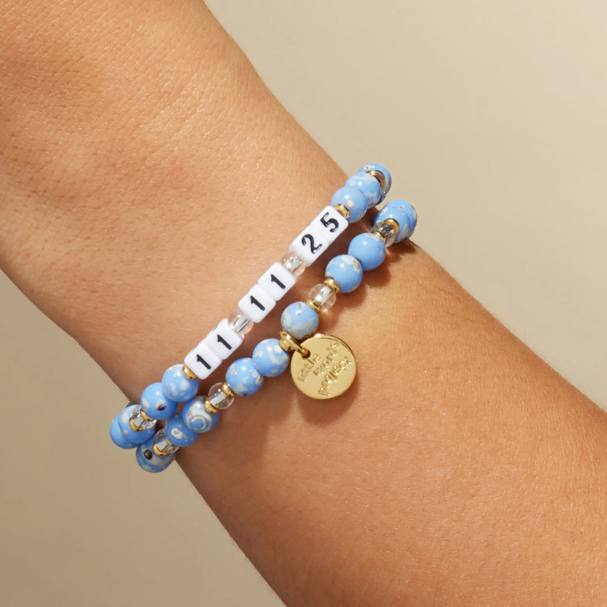 Custom Glacier Glow bracelet with white letter beads reading 11 11 25, light blue speckled and clear crystal beads with a gold LWP charm, on-model on wrist against a tan background