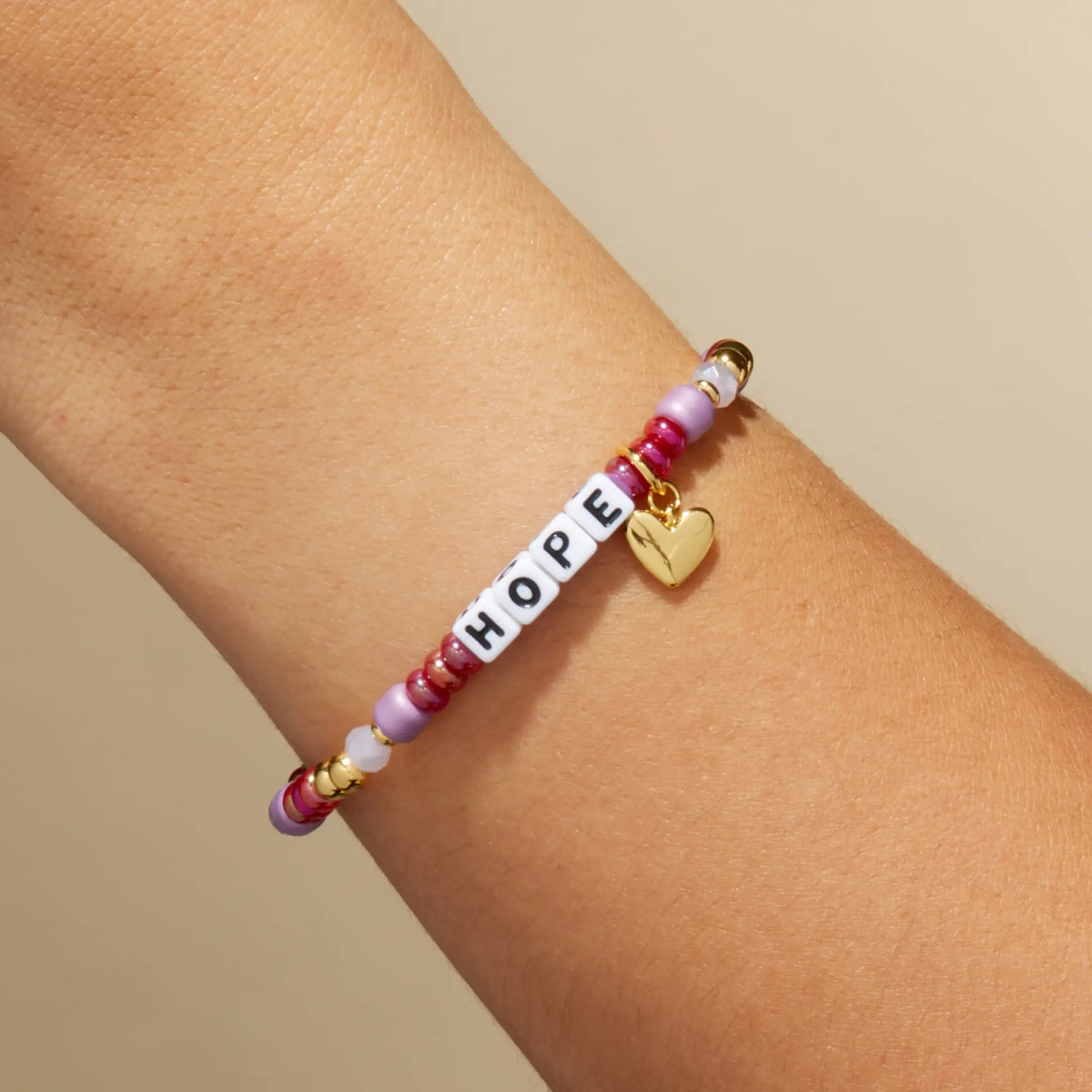 Custom Golden Cherry bracelet with white letter beads reading Hope, cranberry red and mauve beads with a gold heart charm, on-model on wrist against a tan background