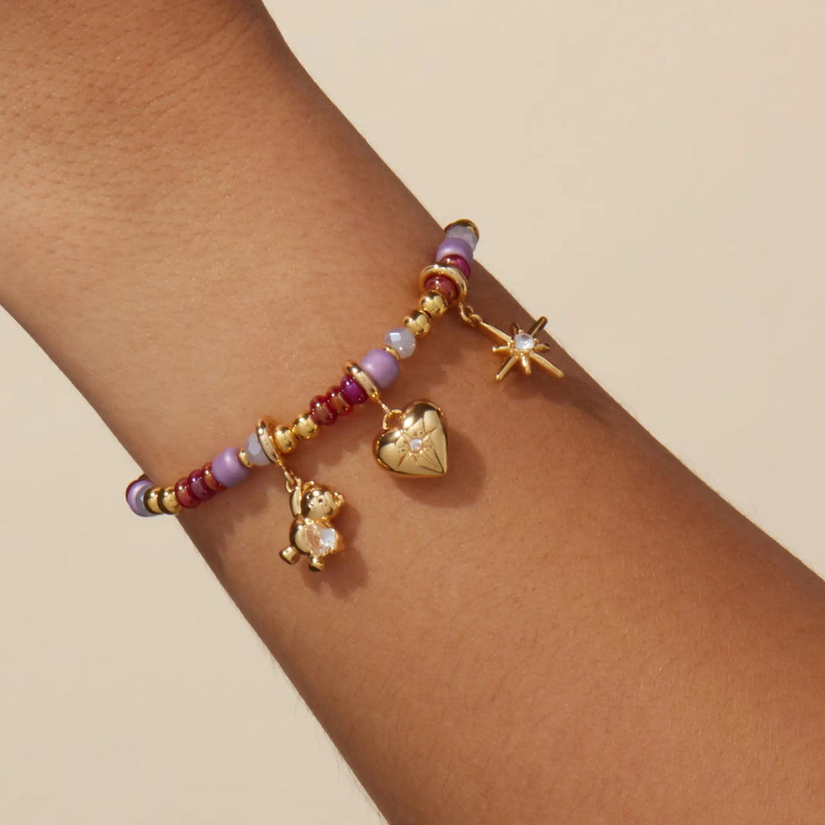 Golden Cherry Charm Bracelet Set worn on a wrist, cherry red, lilac, and gold beads with gold teddy bear gem, cz heart, and starburst charms, on-model on a tan background