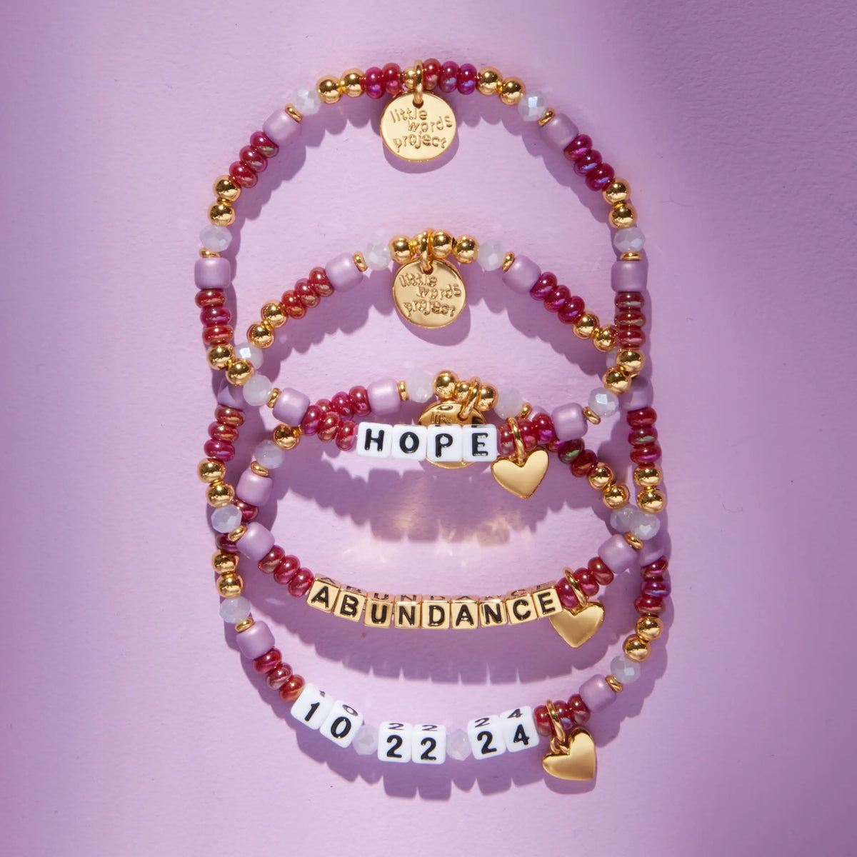 Three Custom Golden Cherry bracelets reading Hope, Abundance, and 10 22 24 in white and gold letter beads, cranberry red and mauve beads with gold heart charms, flat-lay on lavender background
