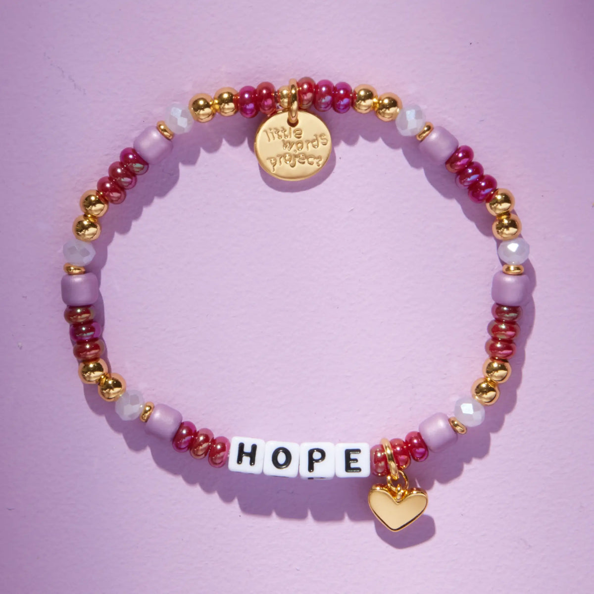 Custom Golden Cherry bracelet with white letter beads reading Hope, cranberry red, mauve, and gold beads with a gold heart charm, flat-lay on lavender background