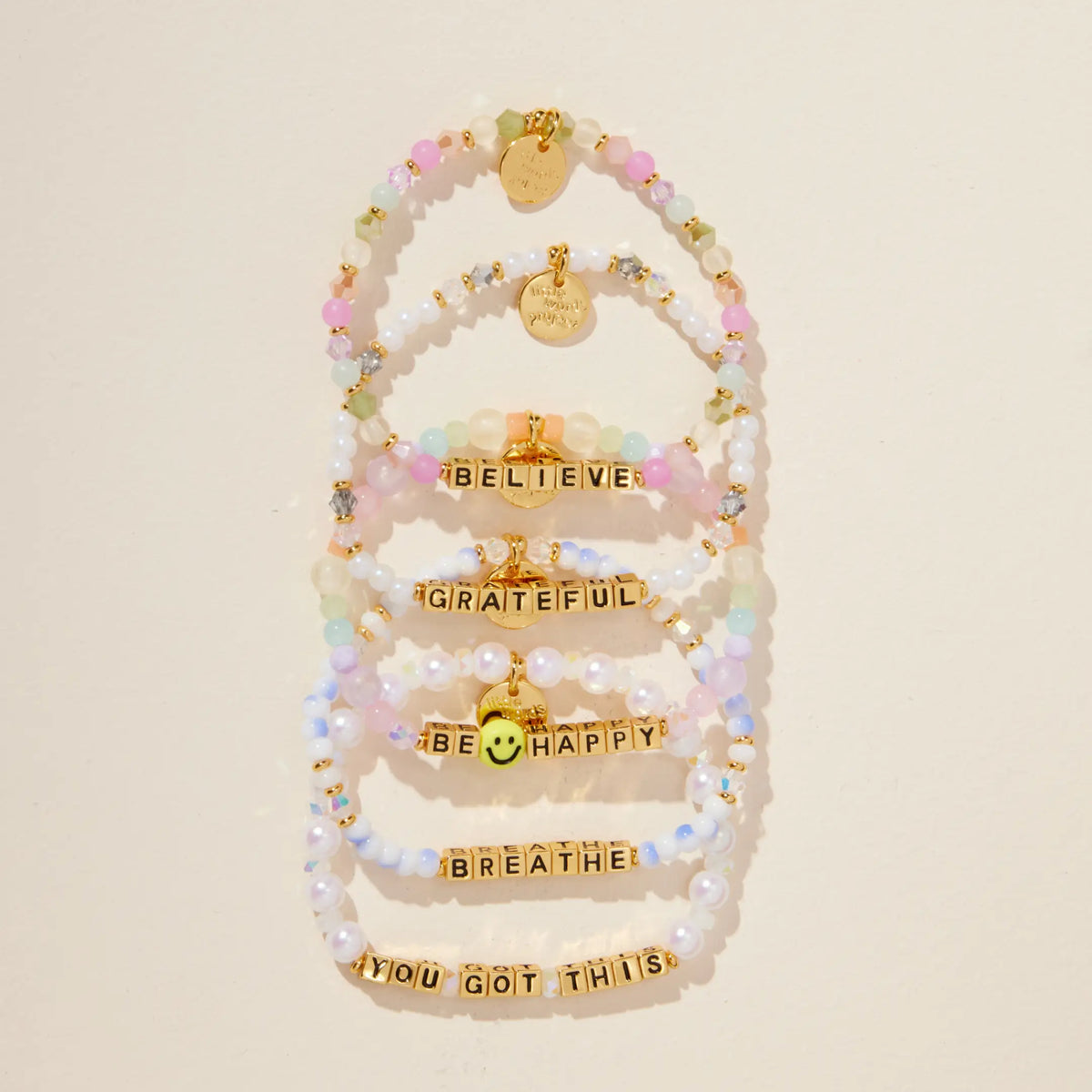 Gold Grateful bracelet in a flat-lay stack of Believe, Grateful, Be Happy, Breathe, and You Got This bracelets with gold letter beads, on cream background