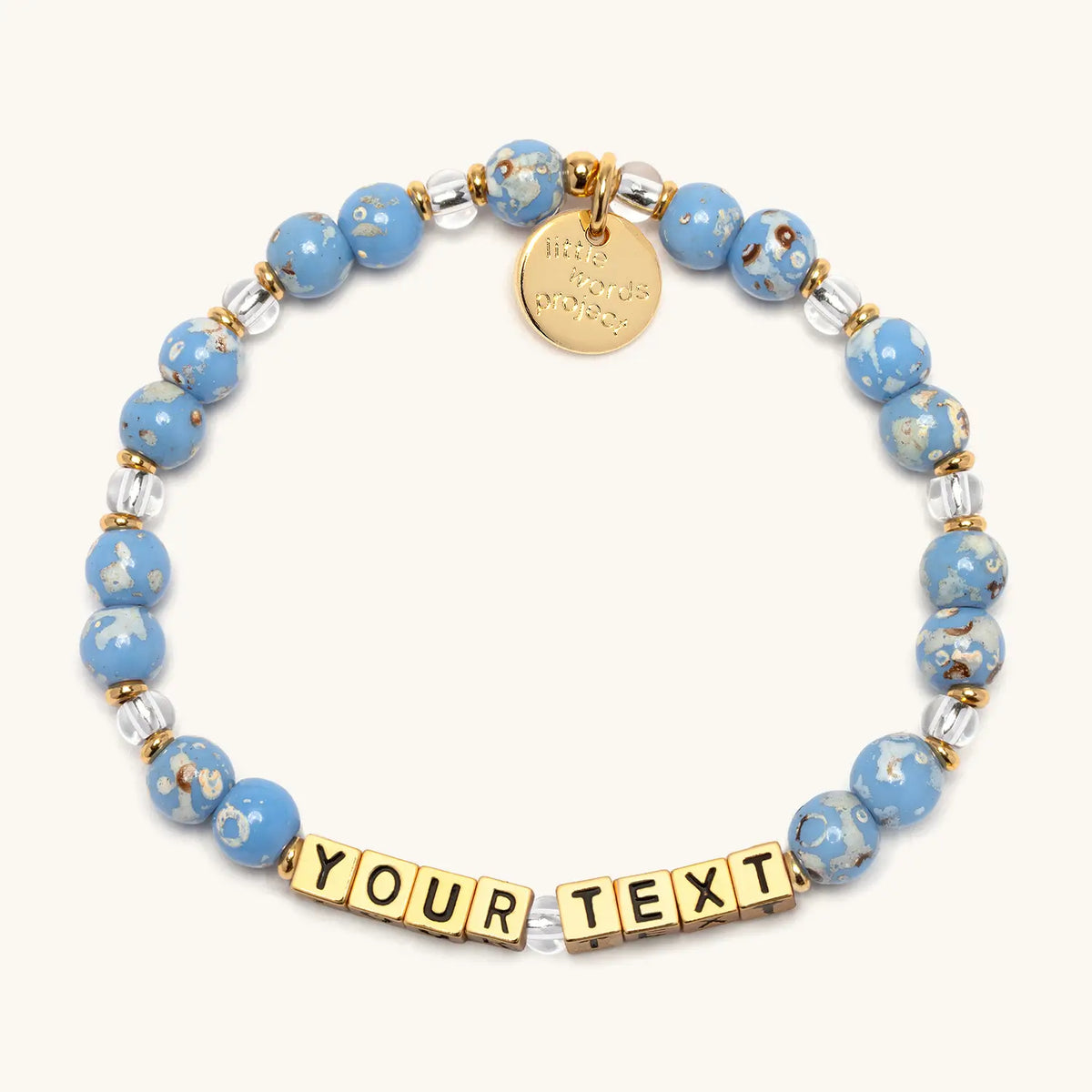 Custom Glacier Glow bracelet in the Glacier Glow bead pattern, gold letter beads reading Your Text, light blue speckled beads and clear crystal beads with gold accents, flat-lay on cream background