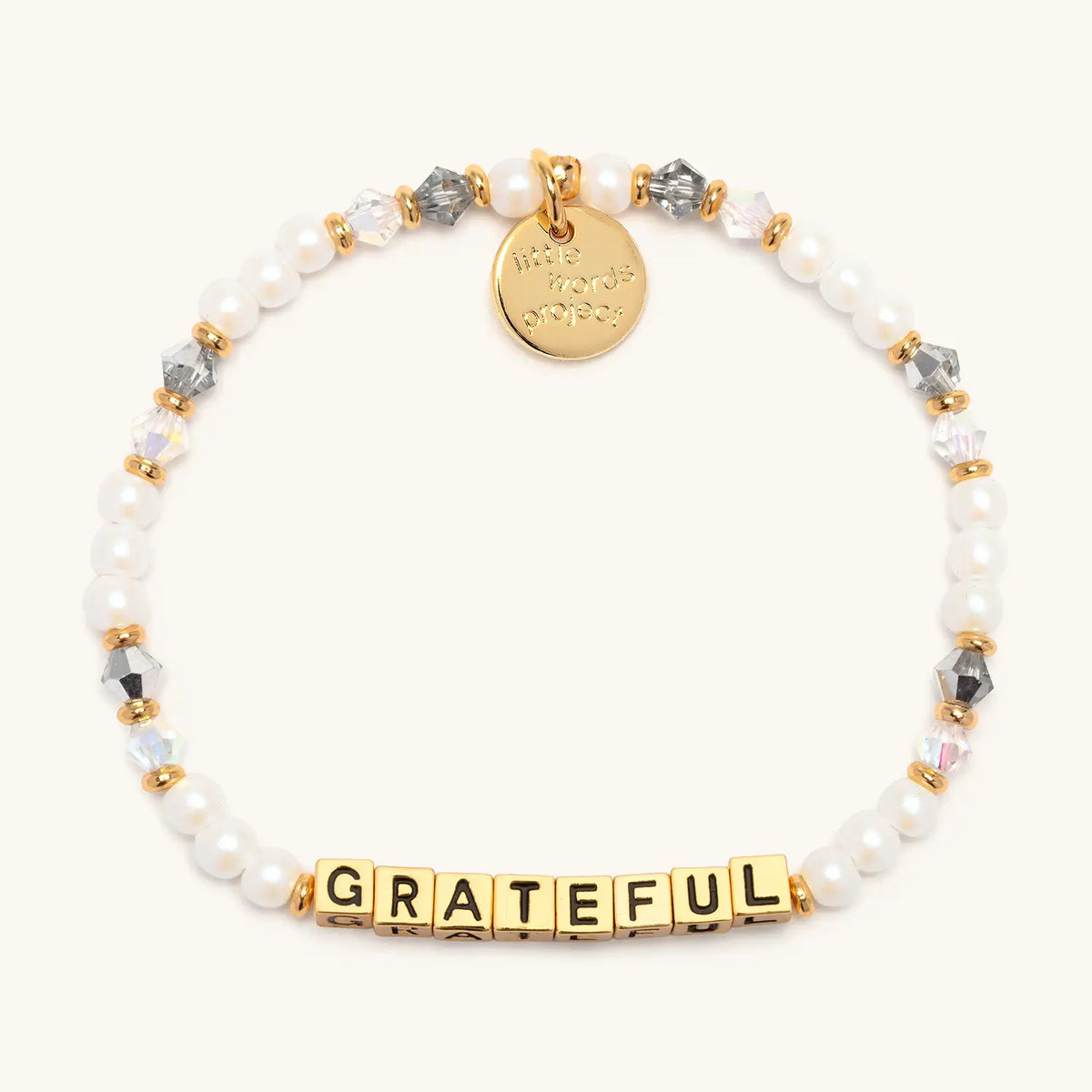 Gold Grateful bracelet in the Shiny Things bead pattern, gold letter beads with white faux pearls and smoky gray and clear faceted crystals with gold accents, flat-lay on cream background