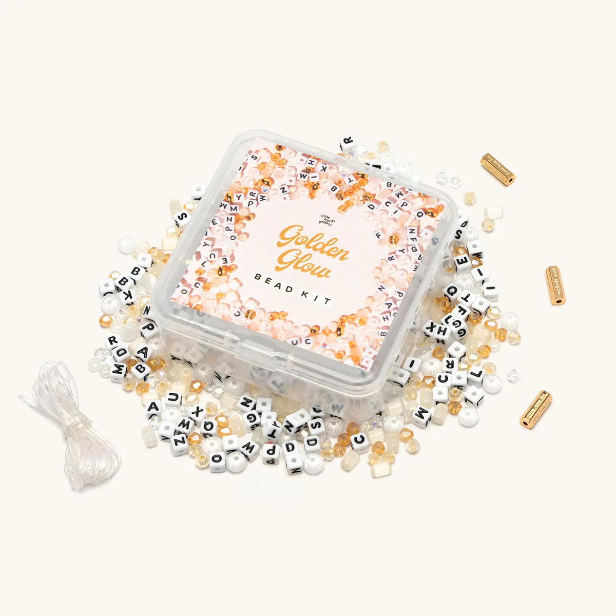 Golden Glow Bead Kit in a clear square tin with a pink and gold label, surrounded by white letter blocks, gold and champagne crystals, white and cream beads, three gold closure beads, and a coil of clear stretch string, flat-lay on cream background