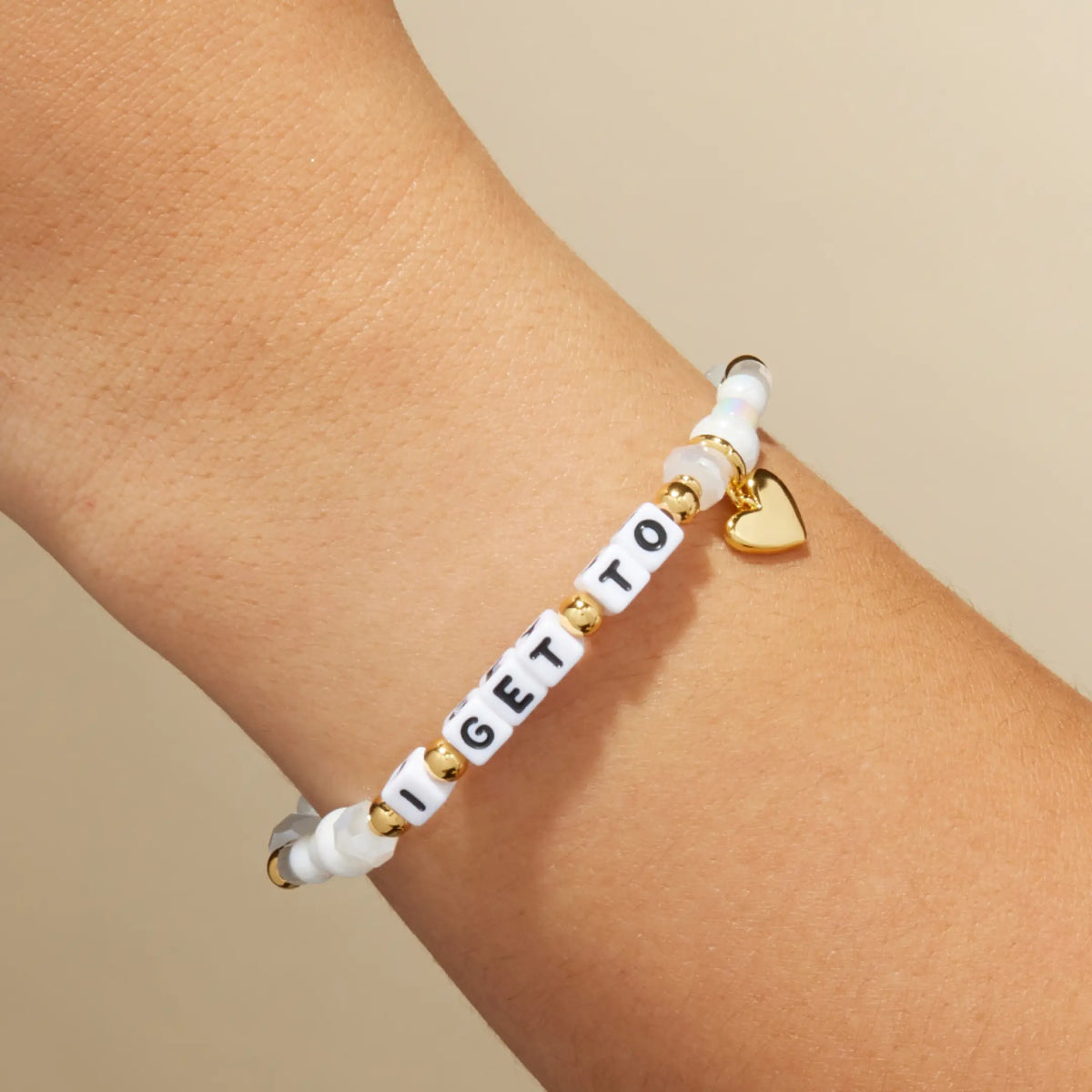 I Get To Charm Bracelet on the wrist, white letter beads with white faux pearls and frosted glass beads, gold accents, and a gold heart charm, on-model on tan background