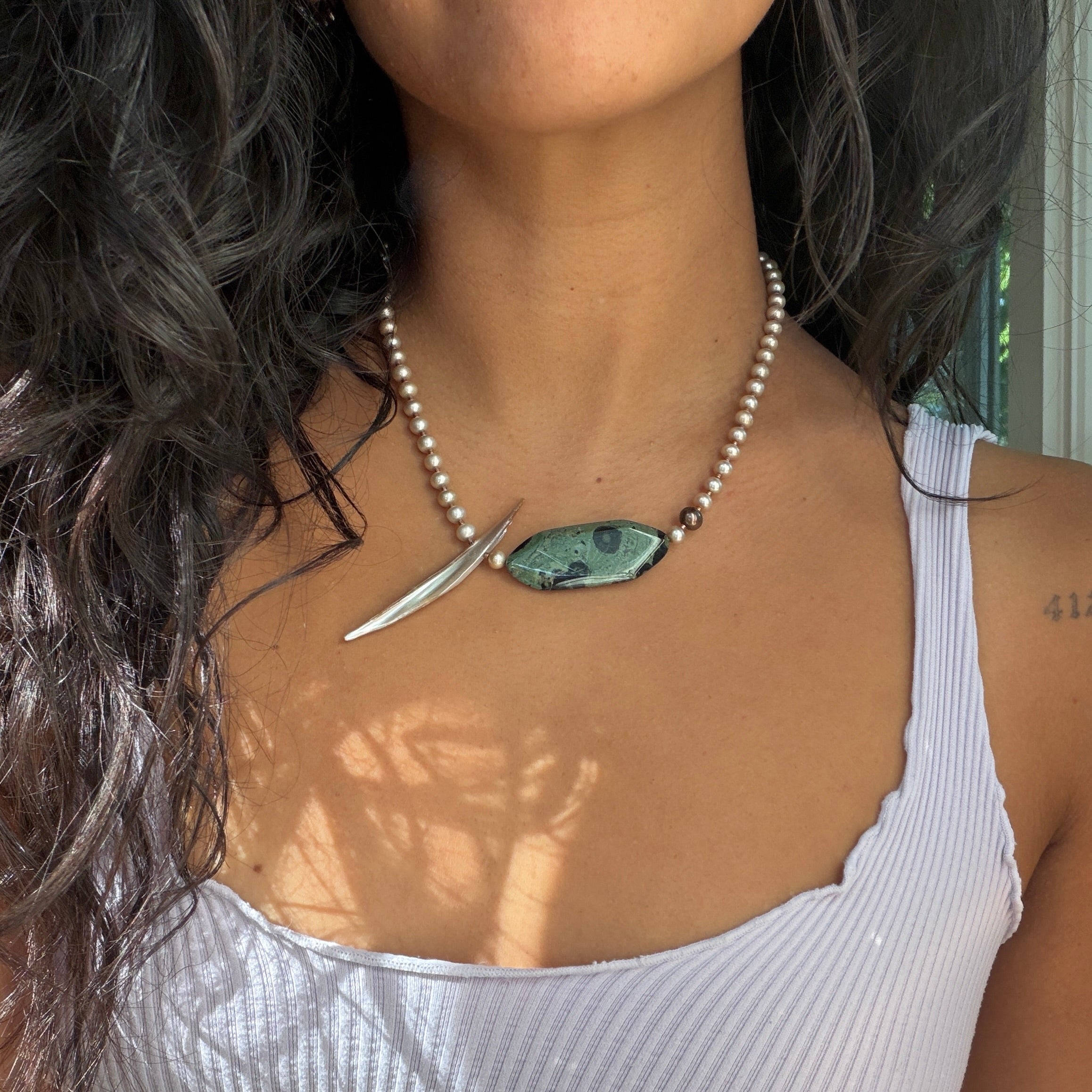 A close-up of a woman wearing a necklace with a silver crescent moon pendant and a green marbled stone pendant, paired with a white ribbed tank top.