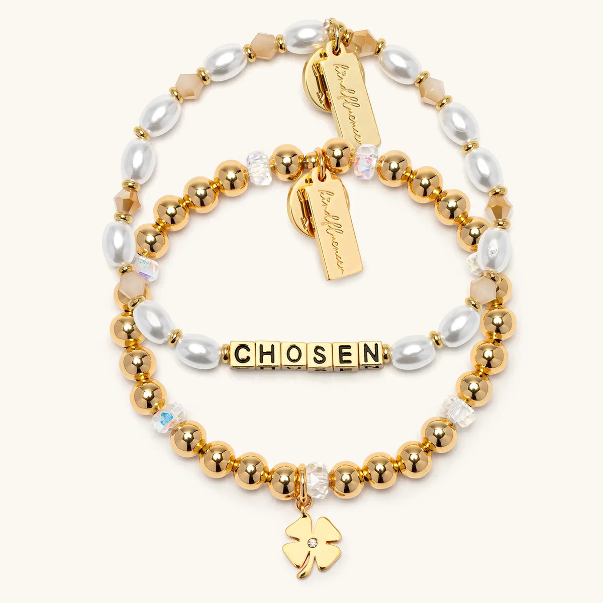 Chosen Bracelet Set by Kirstin Czernek flat-lay on cream background showing two stacked bracelets: the Chosen bracelet in gold letter beads with white faux pearls and champagne crystals, and the Four Leaf Clover Charm Bracelet in gold beads with clear crystals and a gold clover charm