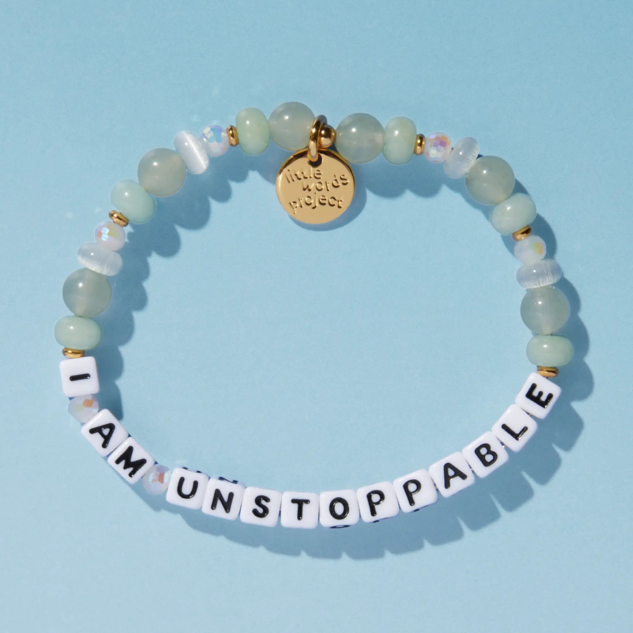A beaded bracelet spelling "I Am Unstoppable" in white letter cubes, with mint green and opalescent beads and a gold Little Words Project charm, on a blue background.