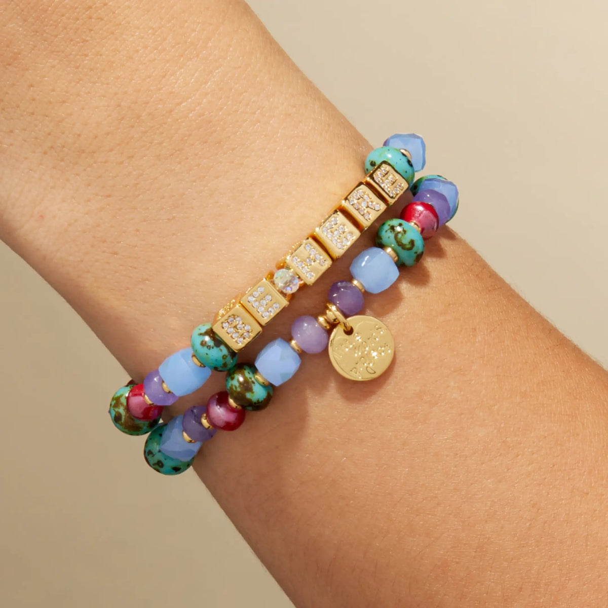 BOLDER Custom Jewel Tones bracelet with gold CZ letter blocks, teal speckled, burgundy, and periwinkle beads, stacked with a second jewel tone beaded bracelet, on-model on wrist against a tan background