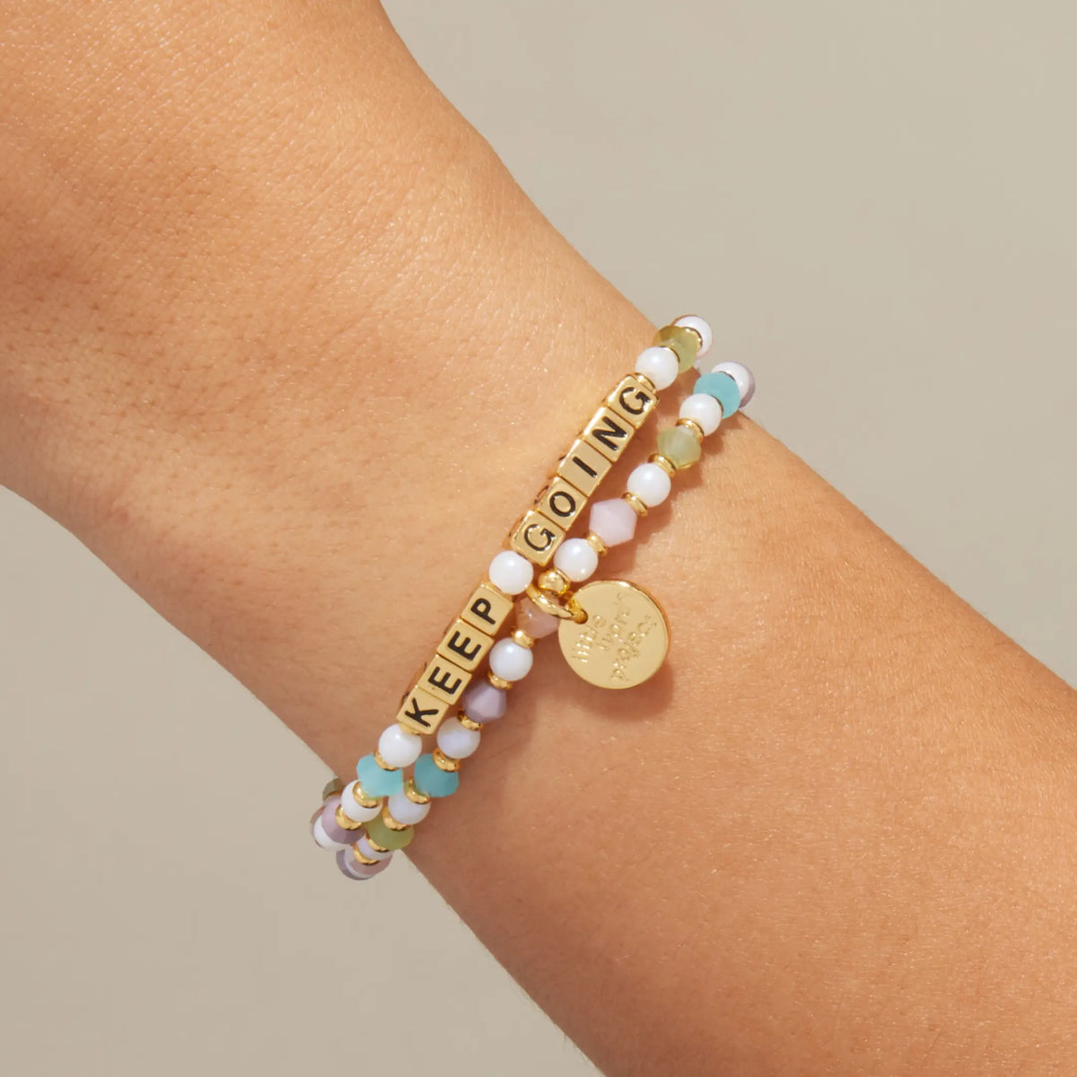 Gold Keep Going bracelet in the Grand Ole bead pattern with gold letter beads, stacked with a coordinating beaded bracelet, on-model on wrist against cream background